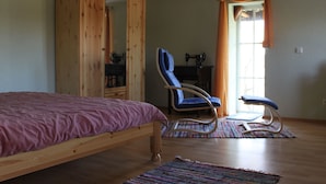 Un dormitorio con un armario de madera, una silla azul, una cama con edredón rosa y una ventana con cortinas naranjas.