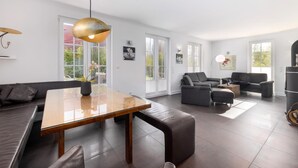 A modern living room with a wooden dining table, leather sofas, and a large window providing natural light. - Orchidee in Glowe by Interhome (Glowe)