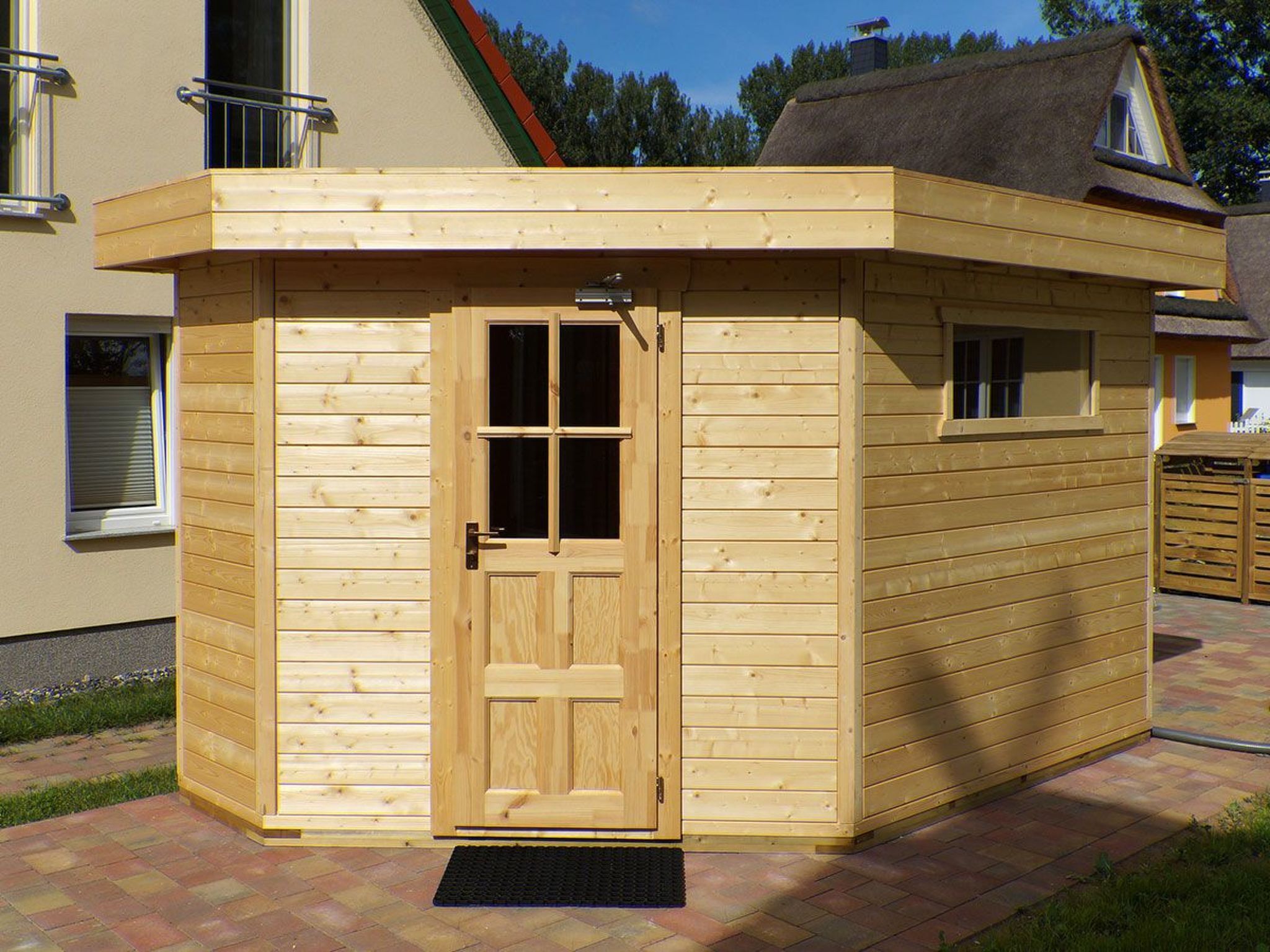 A wooden shed with a door and windows, situated on a paved area with a building in the background.