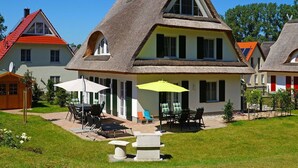Ein traditionelles Haus mit einem steilen Strohdach, einem großen Fenster und einer Terrasse mit Gartenmöbeln.
