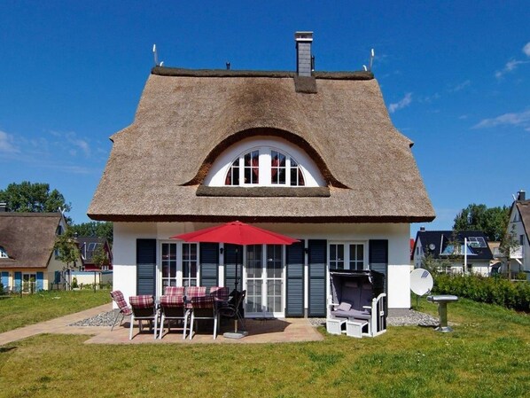 Ein Haus mit einem Strohdach, einer Terrasse mit Gartenmöbeln und einer Sat-Schüssel.