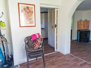 Ein Zimmer mit einem Korbstuhl, einem gerahmten Gemälde, einem Holzschrank und einem durch eine offene Tür einsehbaren Küchenbereich.