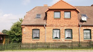 A two-story brick house with a tiled roof, multiple windows with green frames, and a wooden picket fence in front.