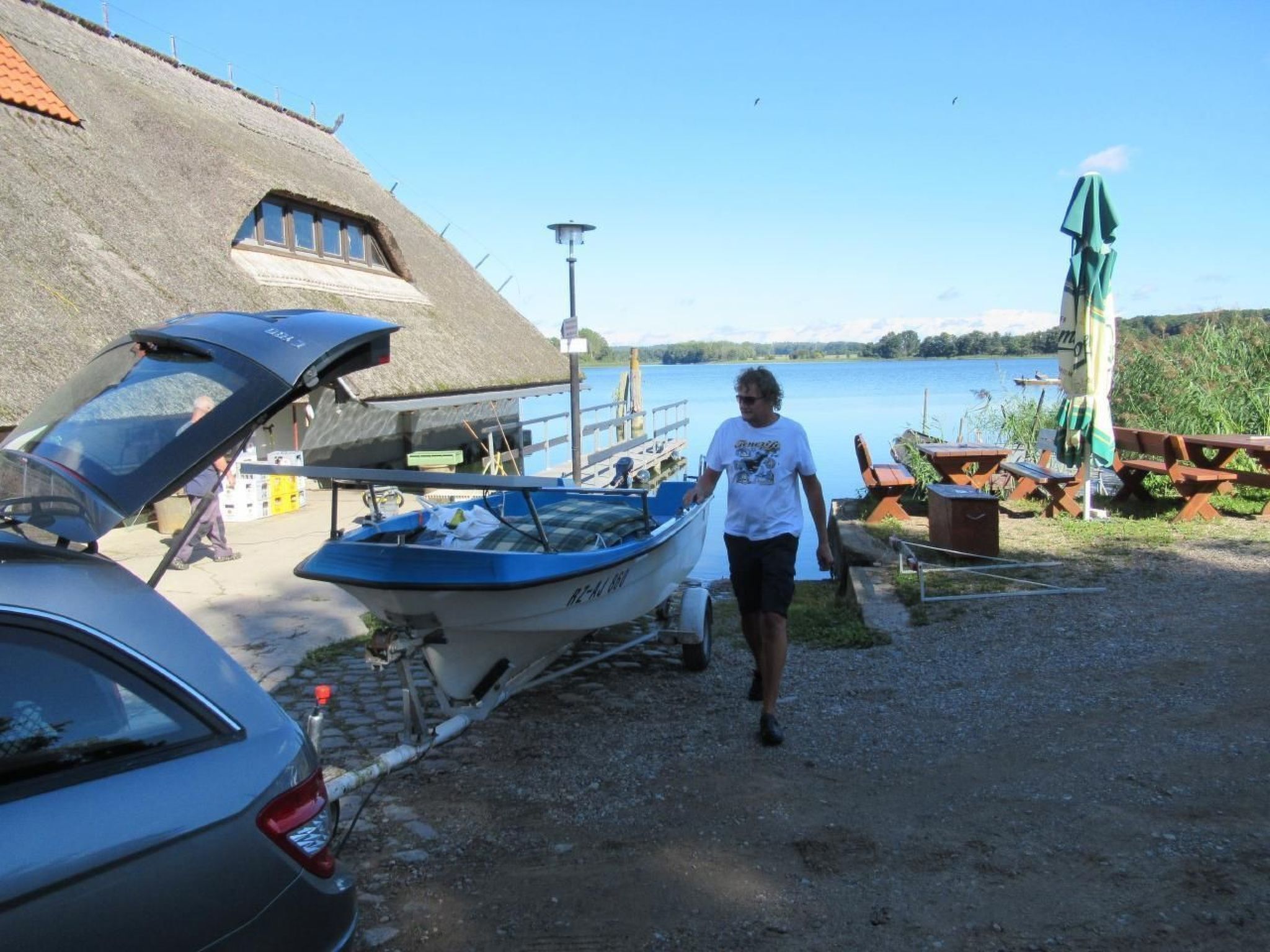 Un bote amarrado cerca de un edificio con techo de paja, un coche y mesas de picnic junto al lago.