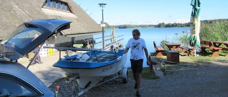 Un bote amarrado cerca de un edificio con techo de paja, un coche y mesas de picnic junto al lago.