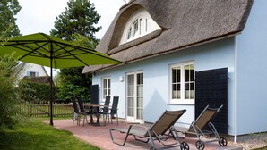 Une maison avec un toit de chaume, une terrasse avec une table et des chaises, et un parasol vert.