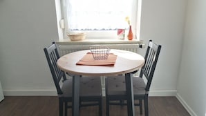 Un comedor con una mesa redonda, cuatro sillas, una cesta, un jarrón con una flor y una ventana con vistas.