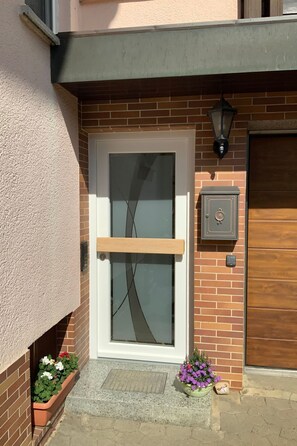 En opgang til boliger med en glasdør, et postkasseanlæg og en lille plantekasse med blomster.