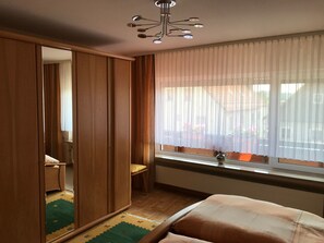 A bedroom with a wooden wardrobe, a bed with a beige cover, a window with sheer curtains, and a ceiling light fixture. - Freistehendes Ferienhaus mit Südbalkon by Interhome (Marktrodach)