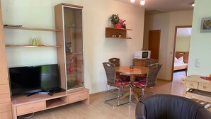 A living room with a flat-screen TV, a glass-fronted cabinet, wicker chairs, a wooden dining table, and a microwave. - Ferienresidenz by Interhome (Bad Kötzting)