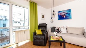 Un salon moderne avec un fauteuil en cuir, un canapé et une table basse. Il y a une grande fenêtre avec des rideaux, un radiateur et un tableau accroché au mur.