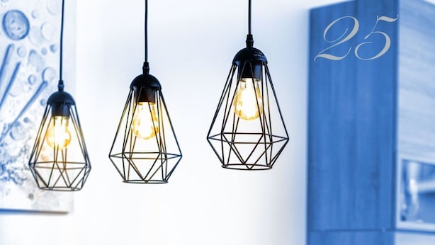 Un coin repas avec une table en bois dressée pour quatre personnes, agrémenté de luminaires suspendus aux formes géométriques, d’orchidées blanches et d’un mur bleu orné du chiffre « 25 ».