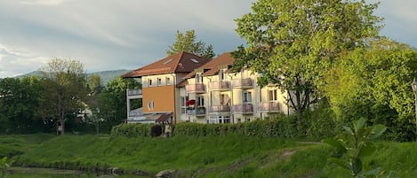A multi-story residential building with balconies, a pond in the foreground, and lush greenery.
