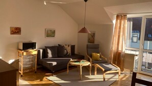 A cozy living room with a sofa, a small table, and a chair. There is a wooden cabinet with a television on top. The room has a sloped ceiling and large windows with curtains. - Ferienresidenz Am weißen Regen by Interhome (Bad Kötzting)
