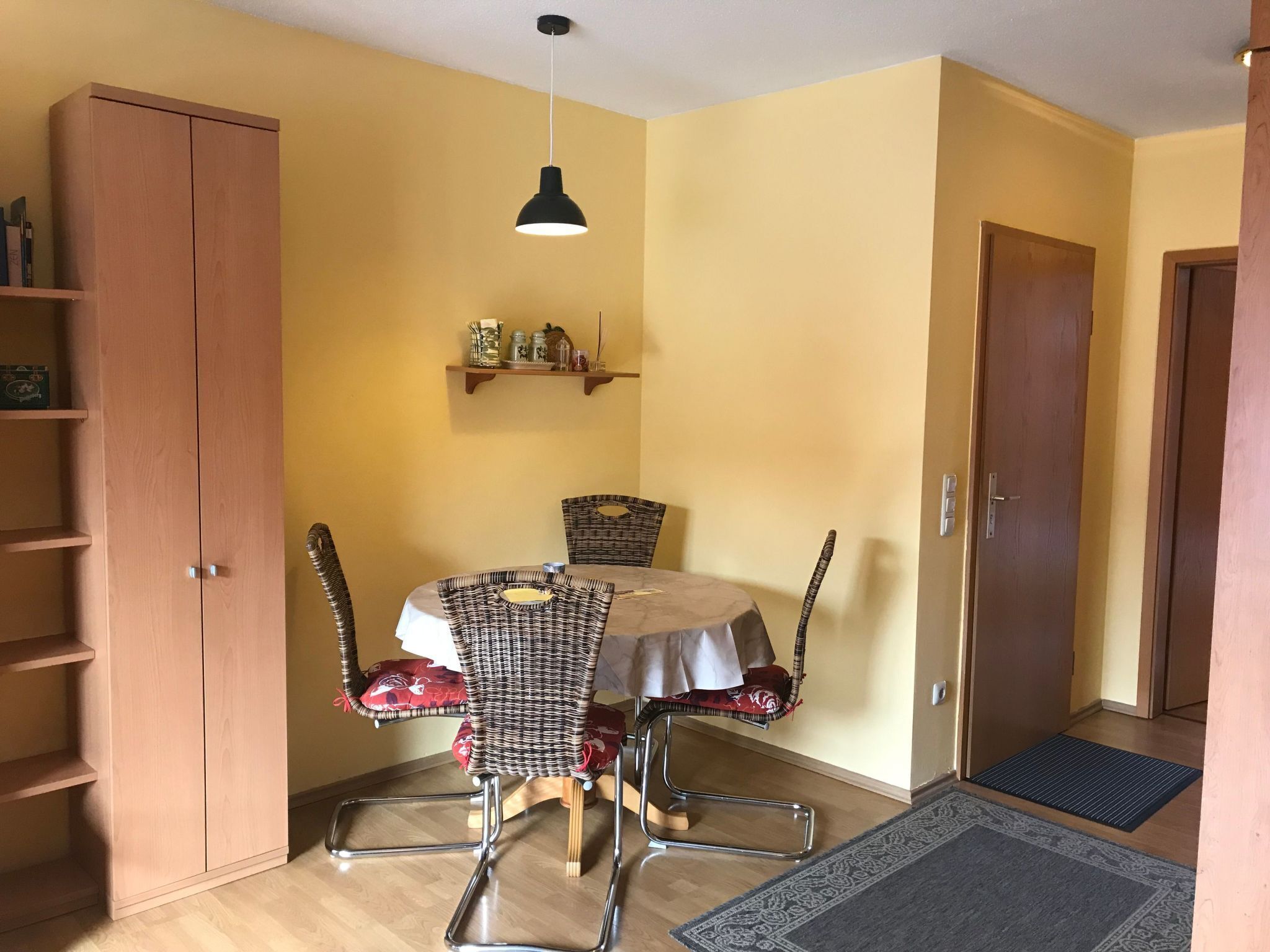 A dining area with a round table, four wicker chairs, a hanging pendant light, a wooden cabinet, and a door.