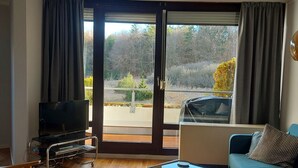 Olohuoneessa on sohva, sohvapöytä ja televisio. Siellä on myös liukuovi, joka johtaa parvekkeelle, jolta on näkymä puita ja autoa kohti.