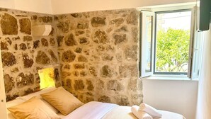 Un dormitorio con pared de piedra, cama con ropa de cama color beige y ventana con vista a la vegetación.