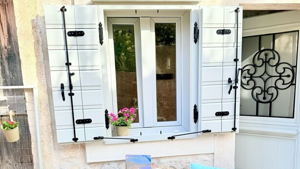 Un pequeño espacio al aire libre con dos sillas y una mesa, una ventana con contraventanas y un portón metálico decorativo.