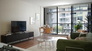 Uma sala de estar moderna com televisão de tela plana, mesa de jantar com cadeiras, sofá verde e grandes janelas com vista para um edifício.