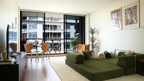 Uma sala de estar moderna com um sofá verde, uma área de jantar com mesa e cadeiras, e grandes janelas com vista para a paisagem urbana.
