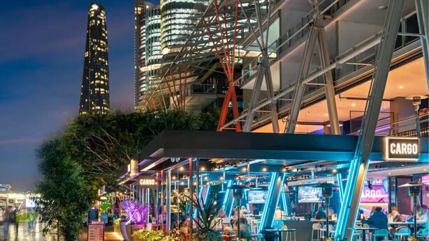 Uma área moderna de refeições ao ar livre com um letreiro 'CARGO', rodeada por vegetação e edifícios urbanos.