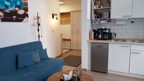 Ett modernt kök med vita skåp, en kaffemaskin och en diskmaskin. Där finns en blå soffa och ett litet matbord med en dekorativ skål.