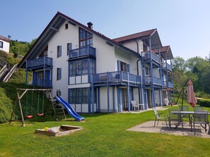 Een huis met twee verdiepingen, met een blauw balkon, een glijbaan en een terras met buitenmeubilair.