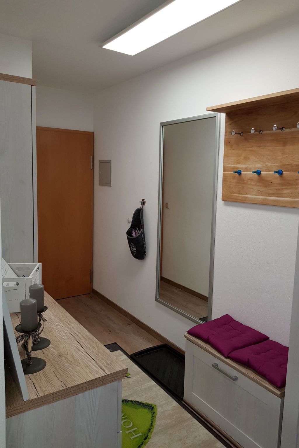 Une petite pièce bien éclairée, avec un plancher en bois, un miroir, une étagère en bois munie de crochets, un coussin violet et une petite poubelle.