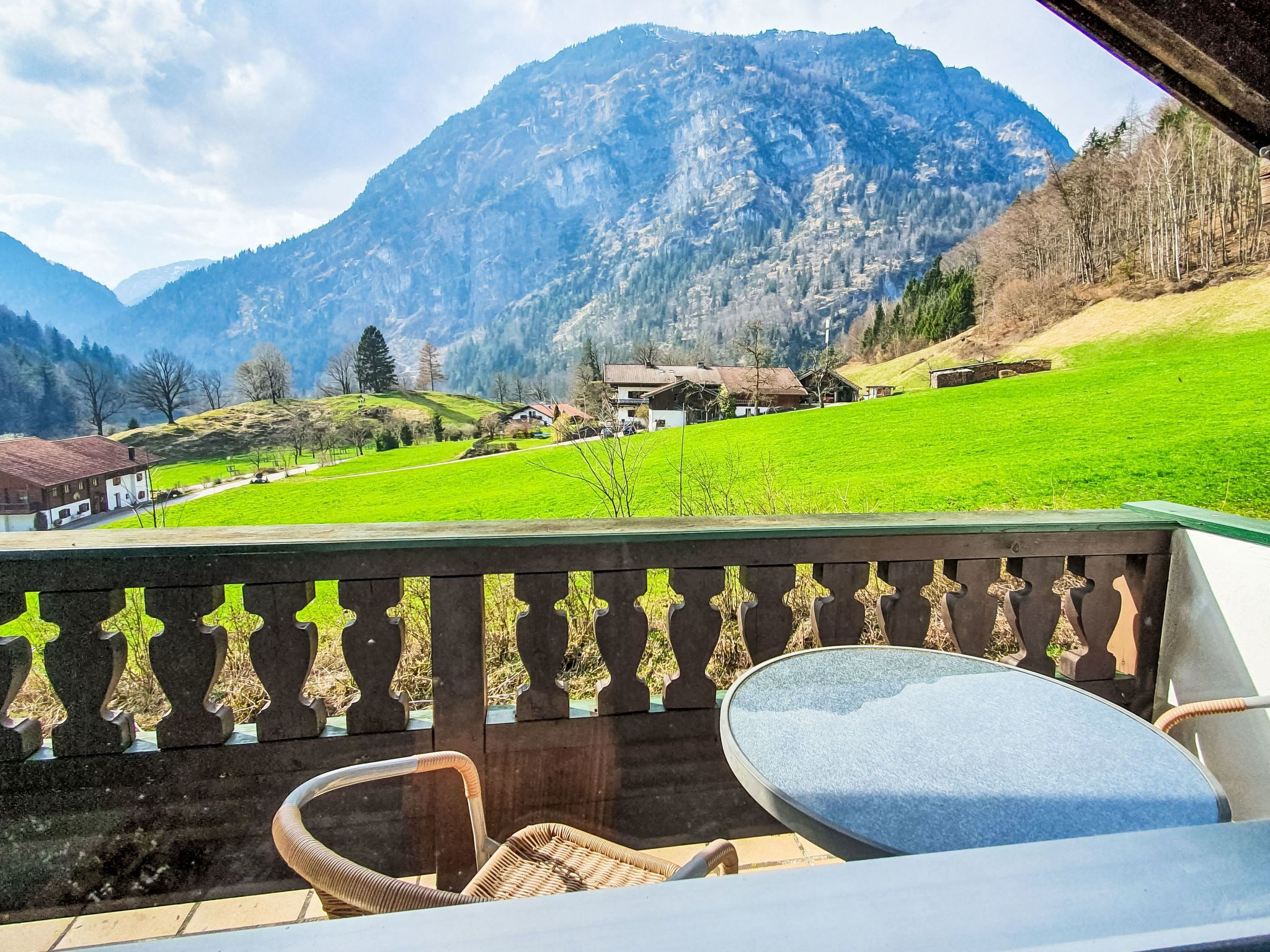 A balcony with wicker chairs and a table, overlooking a green field and houses with mountains in the background.
