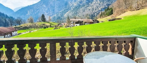 A balcony with wicker chairs and a table, overlooking a green field and houses with mountains in the background.