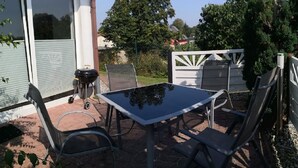 Een terras buiten met een tafel, stoelen, een barbecue en een overdekt gedeelte met een witte schutting.