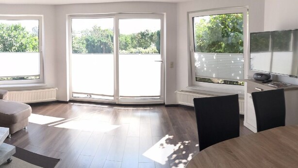 Een moderne woonkamer met een grote schuifdeur naar een balkon, een eethoek met een ronde tafel, en een ingebouwde televisie.