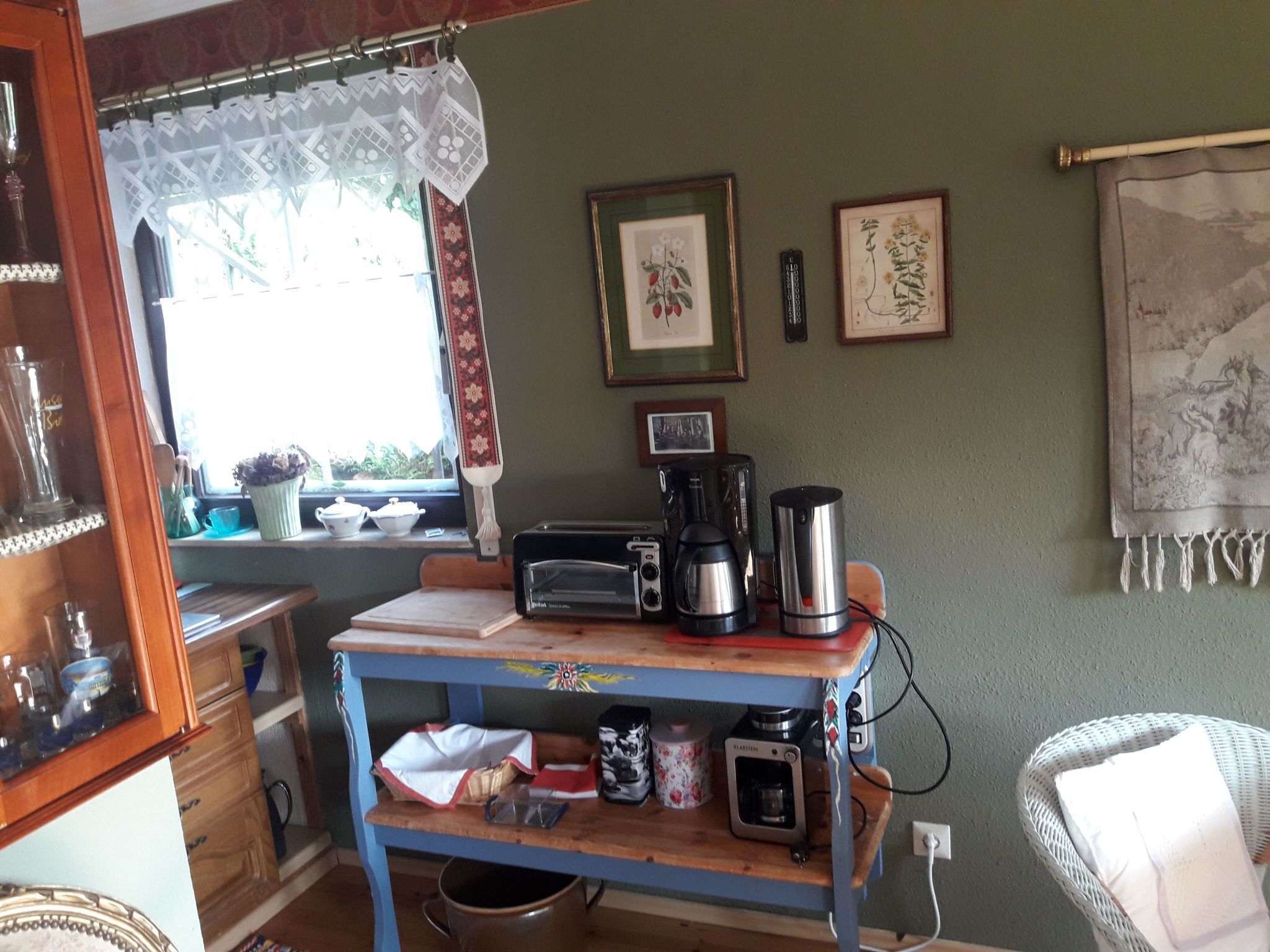 A kitchen with a blue table, a coffee maker, a toaster, and a kettle. There are framed pictures on the wall and a window with curtains.