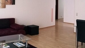 En moderne stue med et glas sofabord, en mørkerød sofa og en sort skammel. Et maleri hænger på væggen, og en dør fører til et andet rum.