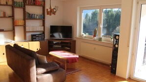 Una sala de estar con un sofá de cuero marrón, un televisor, suelo de madera, una estantería y una ventana con vistas a árboles.