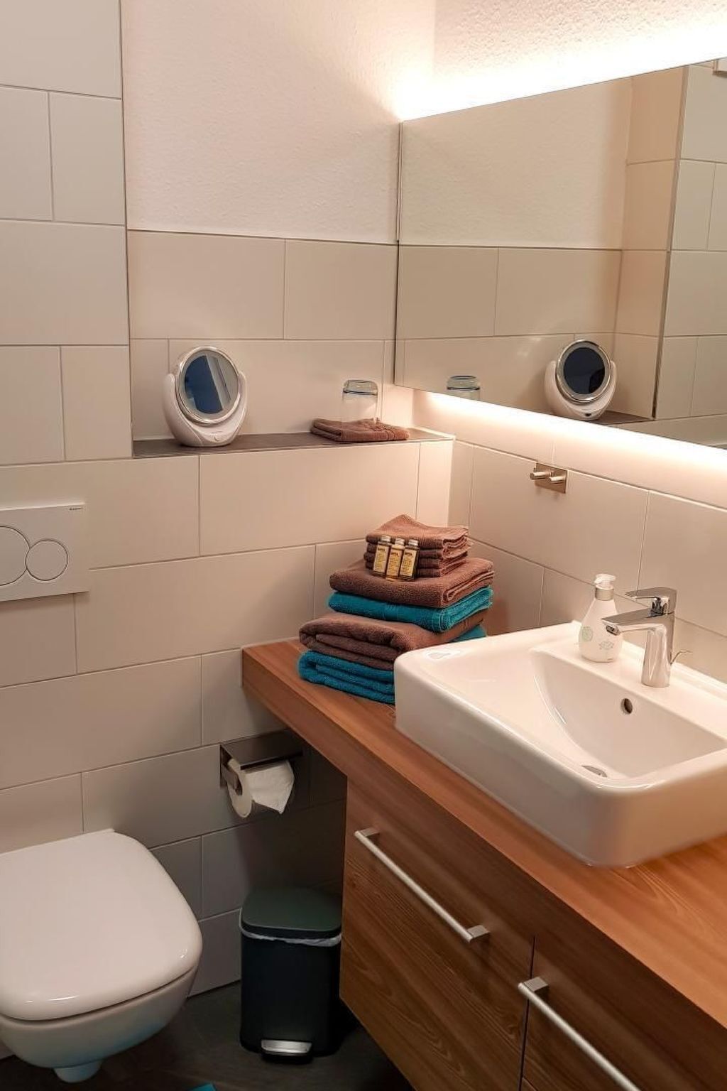 A bathroom with a white sink, mirror, and toilet. There are towels stacked on a wooden shelf, and a trash bin is visible.