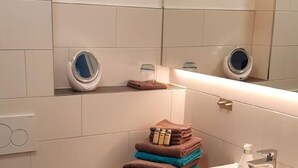 A bathroom with a white sink, mirror, and toilet. There are towels stacked on a wooden shelf, and a trash bin is visible. - Wohnung in Neckarwestheim by Interhome (Neckarwestheim)