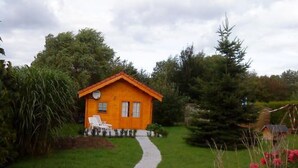 Ein kleines, eingeschossiges Holzhaus mit geneigtem Dach, einem gepflasterten Weg zum Eingang und einem Garten mit verschiedenen Pflanzen sowie einem kleinen weißen Stuhl.
