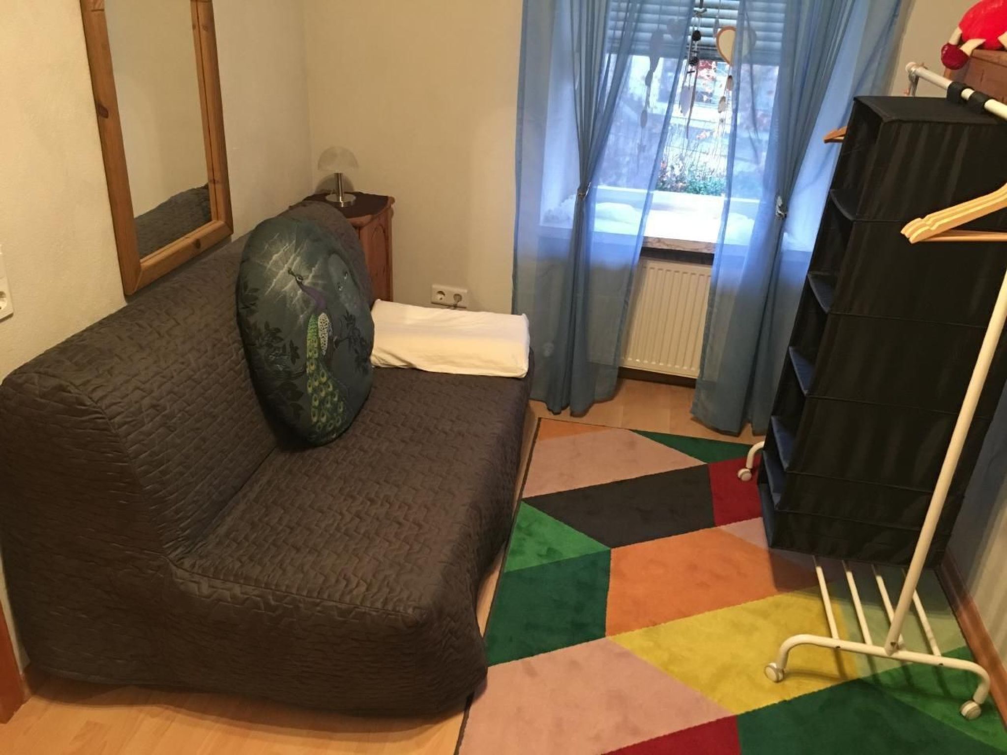 A room with a dark-colored sofa, a small side table with a lamp, a window with blue curtains, and a black storage unit with a white ladder.