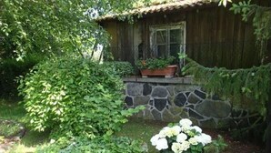 A rustic wooden cabin with a stone wall and a flower bed with white flowers. - in Bernsfeld mit großem Garten by Interhome (Atzenhain)