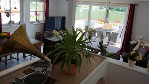 Ein geräumiges Wohnzimmer mit großem Fenster, einem Klavier, einem Fernseher und diversen Pflanzen.