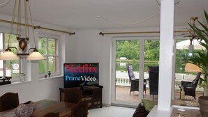 Ein Essbereich mit Fernseher, der 'Netflix Prime Video' zeigt, einem Esstisch, Stühlen und Blick auf einen Garten durch eine Glastür.