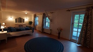Ein Schlafzimmer mit Bett, Nachttisch, Lampe, Kommode und einem großen Fenster mit Vorhängen.