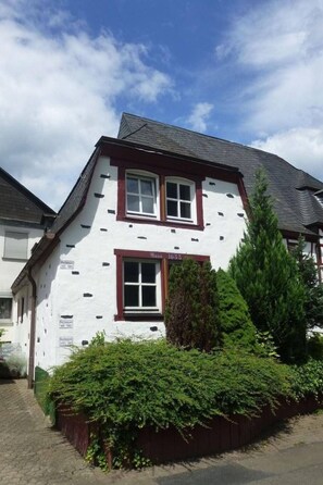 Ein zweistöckiges Gebäude mit weißer Fassade und roter Ornierung. Im oberen Stockwerk befinden sich zwei Fenster, und davor ein kleiner Garten mit grünen Sträuchern.
