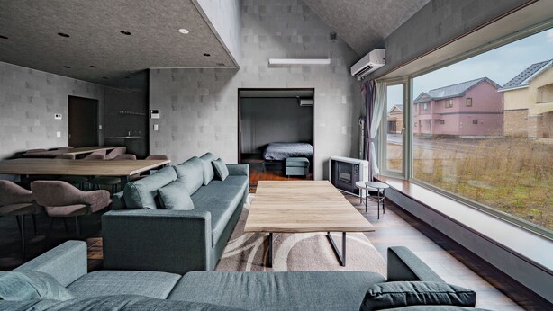 A modern living room with a large window, a sofa, a wooden coffee table, and a dining area with chairs. - PROSTIMO (Bibai)