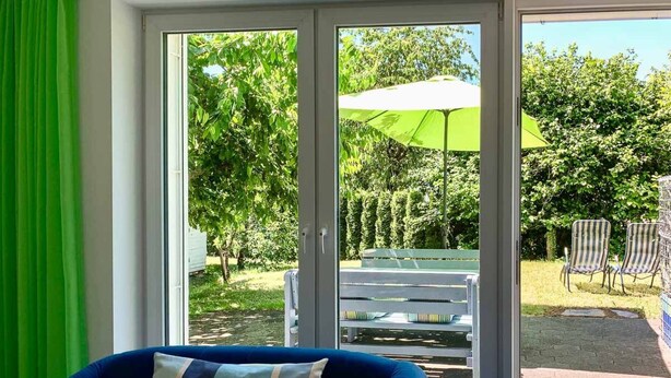 Une pièce avec un canapé bleu et un coussin à rayures, une porte-fenêtre coulissante donnant sur un espace extérieur aménagé avec un banc, des chaises et un parasol vert.