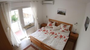 Ein Schlafzimmer mit einem hölzernen Bett, floralem Bettzeug, Blick ins Freie, einem kleinen Tisch mit einer Vase und einem Heizkörper.
