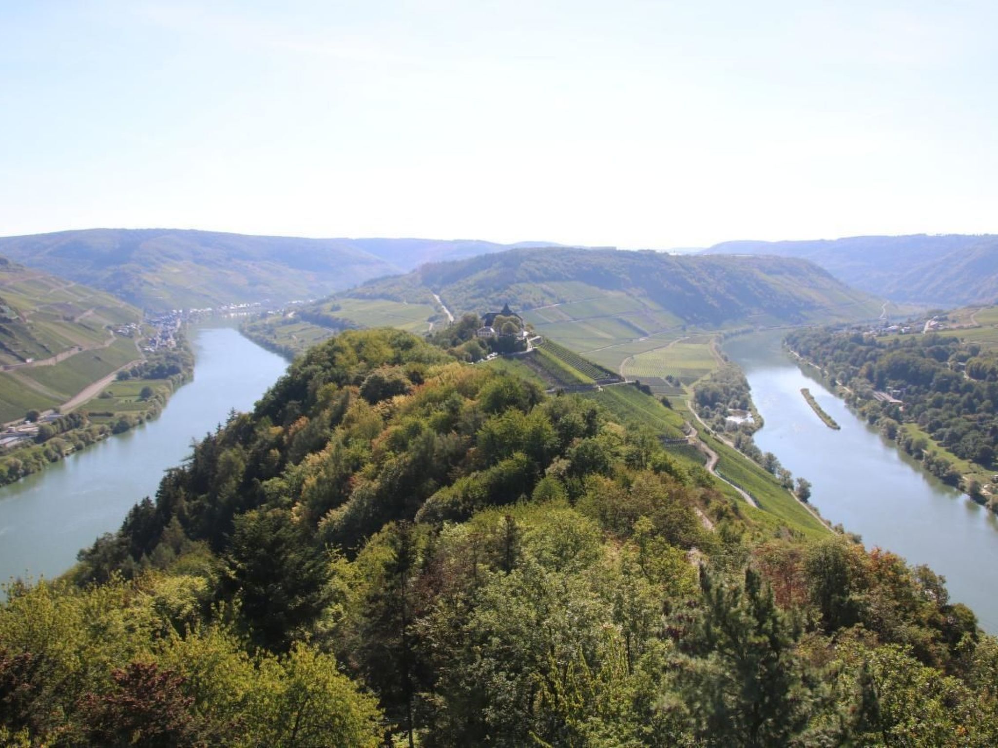 A river valley with a prominent hill in the foreground, surrounded by lush greenery and a small settlement at the base.