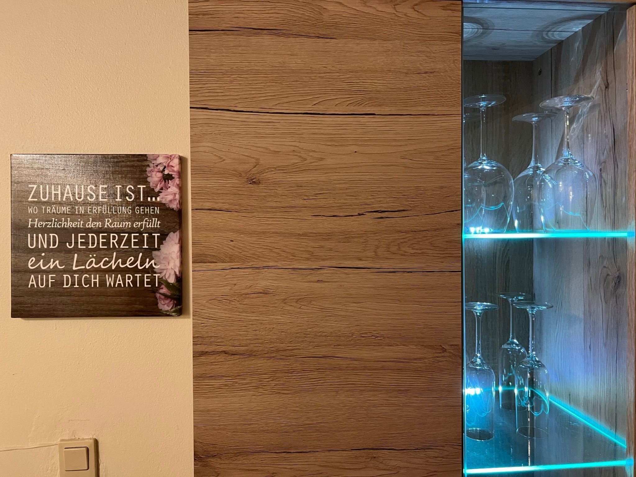 Eine Holzwand mit einem Schild in Deutsch, einem Regal mit Weingläsern und einer Neonleuchte.