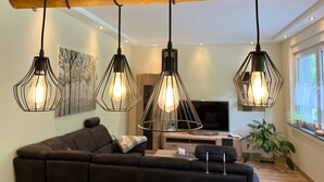 A modern living room with a wooden ceiling beam, a black leather sofa, a TV, and a large window with plants. - Suite am Radweg by Interhome (Pittenbach)